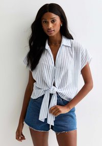 Camisa de manga corta a rayas blancas y azules atada a la cintura, combinada con pantalones cortos de mezclilla. La tela parece ligera y texturizada.