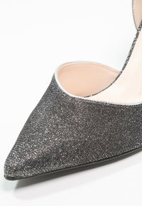 Escarpin à bout pointu en tissu noir texturé avec un éclat métallique. Présente une bordure argentée le long du bord et un intérieur lisse.