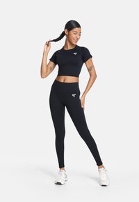 Czarny krótki top z krótkim rękawem oraz legginsy z wysokim stanem, obie rzeczy o gładkiej teksturze i z białym akcentem logo, zestawione z białymi trampkami.
