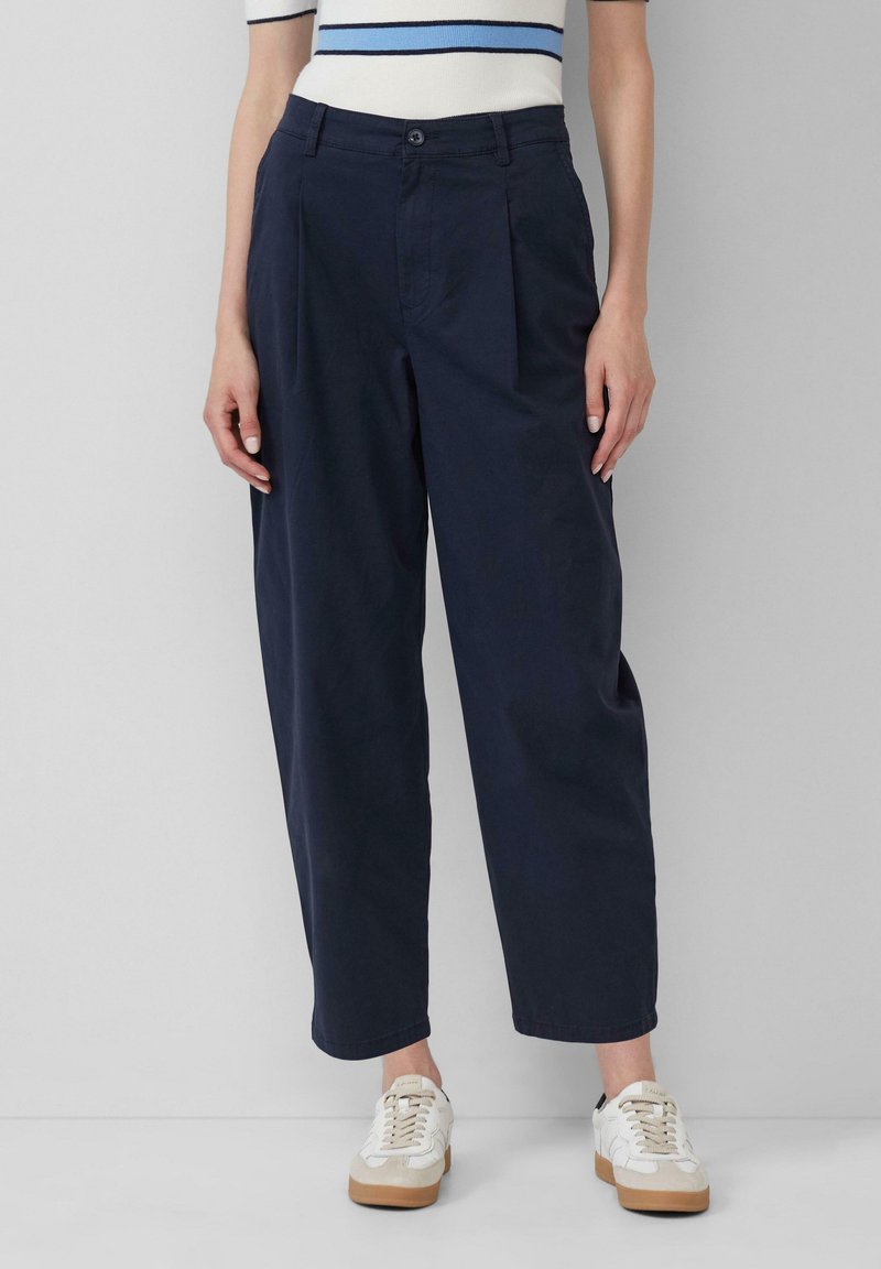 Pantaloni a gamba larga blu navy con pieghe frontali, realizzati in cotone, con chiusura a bottone e tasche laterali, abbinati a sneakers bianche.