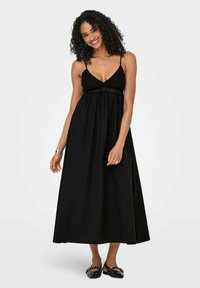 Zwarte maxi-jurk met een aansluitend bovenlijfje, voorzien van dunne spaghettibandjes, textuurstof en een vloeiende rok. Draagt met zwarte platte schoenen.