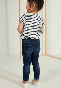 Otrok, oblečen v kratko majico s črtami in temno modre skinny jeans, s hrbtnimi žepi in belimi čevlji s kovinskimi poudarki.