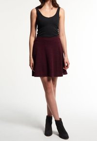 Femme portant un haut sans manches noir, une jupe évasée rouge foncé et des bottes noires à la cheville, se tenant les jambes croisées devant un fond blanc.