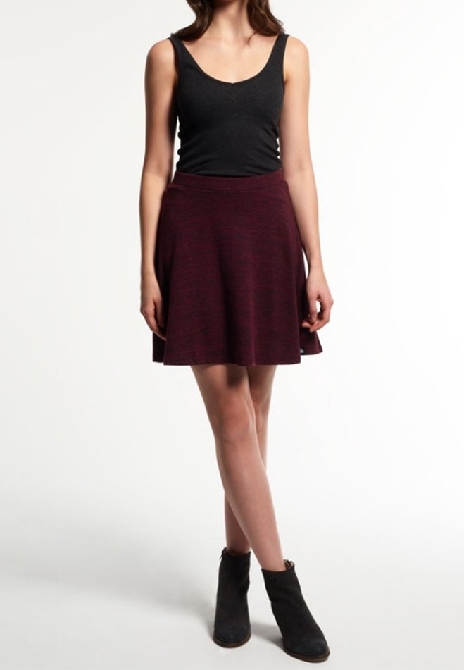 Femme portant un haut sans manches noir, une jupe évasée rouge foncé et des bottes noires à la cheville, se tenant les jambes croisées devant un fond blanc.