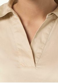Camisa beige con cuello de pico, textura suave y detalles de costura visibles cerca del cuello y a lo largo del frente.