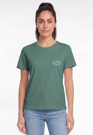 Frau mit dunklen Haaren im Pferdeschwanz, die ein grünes T-Shirt mit weißem deutschen Text und hellblaue Jeans trägt, steht vor einem weißen Hintergrund.
