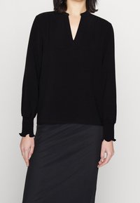 Mustapaita, jossa on pitkät hihat ja V-pääntie, varustettu rypytetyillä hihansuilla ja pehmeällä, tekstuurilla kankaalla. Yhdistetty sileään mustaan hameeseen.