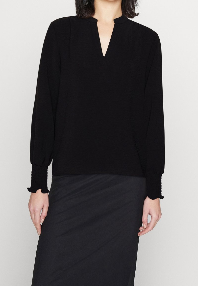 Mustapaita, jossa on pitkät hihat ja V-pääntie, varustettu rypytetyillä hihansuilla ja pehmeällä, tekstuurilla kankaalla. Yhdistetty sileään mustaan hameeseen.