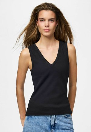 Vrouw met lang bruin haar, gekleed in een mouwloze zwarte V-hals top en lichtblauwe jeans, staande tegen een effen witte achtergrond.