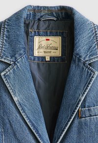 Denim blejzer z reverjem, modre barve in kontrastnim rumenim šivanjem. Notranjost ima gladko tkaninsko podlogo in etiketo. Velikost S.