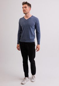 Blauer V-Ausschnitt-Pullover aus weichem Strickmaterial, kombiniert mit schwarzen Hosen und weißen Sneakern. Das Model steht vor einem hellen Hintergrund.