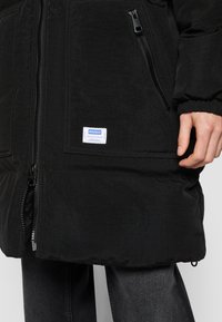 Sort polstret jakke med tekstureret overflade, der har en lynlås foran, sidelommer og et hvidt mærke med blå tekst. Ærmerne er med manchetter.