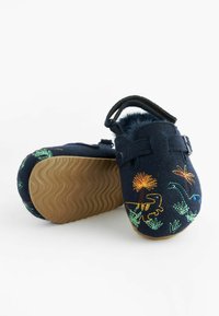 Sabots en feutre navy avec une broderie de dinosaures colorés, une doublure en peluche et une semelle en caoutchouc marron à texture ondulée. Détail de sangle réglable.