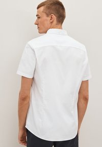 Camisa blanca de manga corta con cuello abotonado, textura lisa y dobladillo curvado. Presenta un yugo trasero con un detalle de botón único.