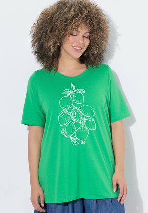 Kvinde med krøllet hår iført en knaldgrøn t-shirt med en hvid stregtegning af citroner og blade, stående mod en hvid baggrund.