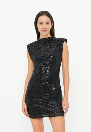 Joven mujer con un vestido mini negro sin mangas con lentejuelas, de pie y sonriendo frente a un fondo blanco liso.