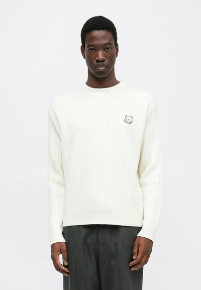 Man wearing a white ribbed sweater with a small wolf logo on the chest and gray trousers against a plain white background.