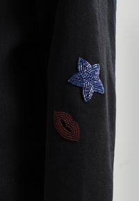 Czarny top z długim rękawem, z teksturowaną niebieską gwiazdą i aplikacją czerwonej wargi na lewym rękawie, wykonany z miękkiej bawełny.