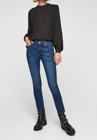 Kvinna iförd en genomskinlig svart blus med lång ärm, blå smala jeans och svarta klackiga boots med nitar, stående med en hand i fickan.