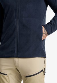 Navy-Fleece-Jacke mit durchgehendem Reißverschluss, elastischen Bündchen und Seitentaschen, getragen über hellbeigen Hosen mit Reißverschlussakzenten.