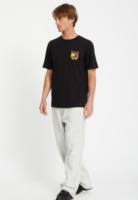 Sort bomulds t-shirt med korte ærmer og et frontgrafik med farverig tekst og elementer. Parret med lys grå jeans og sorte sko.