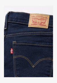Vaqueros de mezclilla azul oscuro con una etiqueta de cuero marrón claro que presenta texto y diseño impresos en rojo. Incluye el logo rojo de Levi's en el bolsillo trasero.