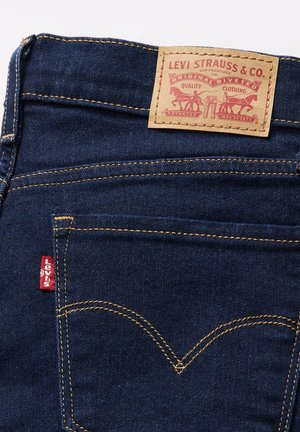 Jeans in denim blu scuro con un'etichetta in pelle color cuoio che presenta testo e design stampati in rosso. Include il logo rosso di Levi's sulla tasca posteriore.
