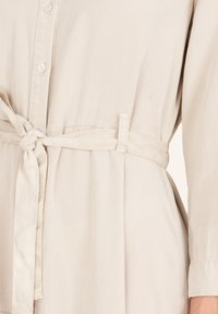 Jasnobeżowa sukienka koszulowa z zapinanym przodem na guziki, paskiem w talii i długimi rękawami. Gładki, lekki materiał o subtelnej teksturze.
