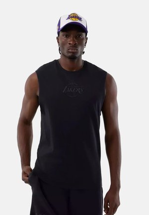 Homme portant un débardeur noir des Los Angeles Lakers et une casquette blanche et violette des Lakers, debout devant un fond uni.