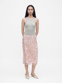 Femme aux longs cheveux foncés portant un haut gris sans manches, une jupe midi rose à sequins et des ballerines beiges pointues, debout sur un fond blanc.