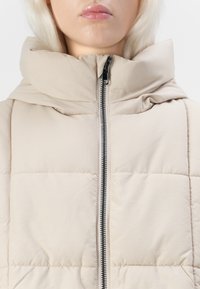 Beige polsterdatud jakk kõrge kraega, millel on täispikk esi-zipper ja tikanditule. Sile kangas, millel on peen sära.