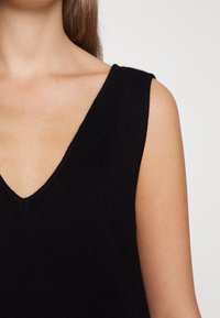 Haut sans manches en tricot noir avec un décolleté en V profond, présentant une texture lisse et des bords nets. La visualisation se concentre sur le décolleté et la zone des épaules.