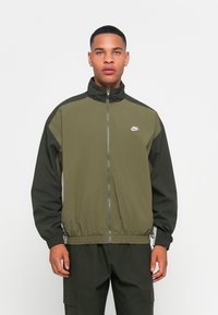 Oliveno zipp-up jopico z visokim ovratnikom, črne poudarke na ramenskem delu in elastične manšete. Na prsih je bel Nike logo.