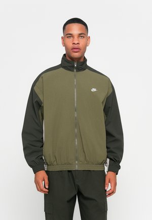 Olive green zip-up jacket with a high collar, black accents on shoulders, and elastic cuffs. Features a white Nike logo on the chest.