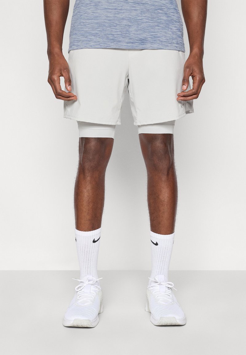 Αθλητικό σορτς ανοιχτού γκρι χρώματος με πλαϊνές τσέπες, σε συνδυασμό με λευκά αθλητικά παπούτσια και κάλτσες με μαύρο λογότυπο. Το πάνω μέρος είναι από μπλε ύφασμα με μελανζέ.