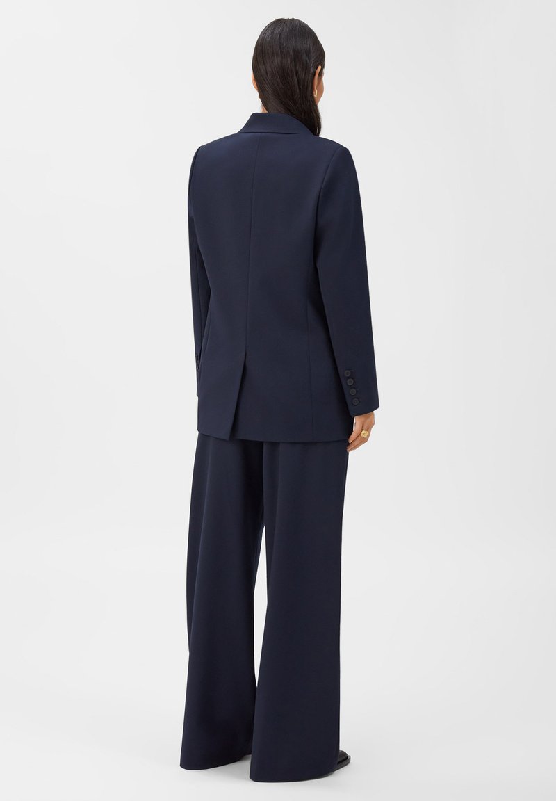Marineblazer und weit geschnittene Hose aus maßgeschneideter Stoffe. Der Blazer hat einen Reverskragen und drei Knöpfe an jedem Ärmelsaum. Glatte Textur.