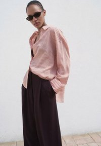 Chemise à manches longues rose avec de fines rayures blanches, associée à un pantalon large noir. Tissu lisse et coupe décontractée.