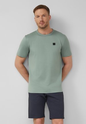 Mann mit kurzen Haaren, der ein salbegrünes kurzärmeliges Hemd und marineblaue Shorts trägt und vor einer hellgrauen Wand steht.