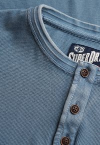 Close-up van de kraag van een blauw getextureerd henley-shirt met drie bruine knopen en zichtbaar Superdry-merklabel.