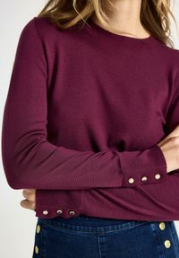 Vrouw in een paarse trui met lange mouwen, voorzien van zilveren knopen op de manchetten en onderrand, gecombineerd met donkerblauwe jeans met gouden knoppen.