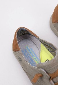 Zapato de lona gris con acentos de ante marrón, que presenta un interior texturizado y una plantilla amarilla brillante. Diseño de punta redonda y cordones.