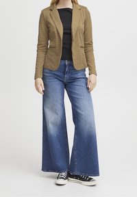 Veston à motif pied-de-poule marron, coupe ajustée, top noir en dessous, jean bleu délavé à jambes larges, et baskets noires avec semelles blanches.