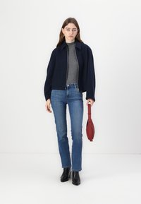 Giacca cropped blu navy con colletto alto, tessuto testurizzato, dolcevita grigia sotto, jeans blu e stivaletti neri alla caviglia. Tiene in mano una borsa rossa.