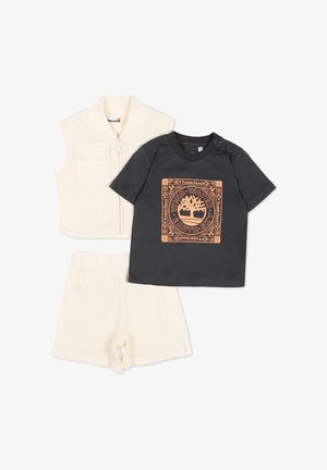 Ensemble trois pièces pour tout-petit comprenant un gilet en maille crème et un short assorti, ainsi qu'une chemise noire à manches courtes arborant un logo Timberland orange et un motif inspiré de la nature.