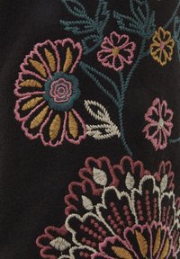 Farverig blomsterbroderi med pink, sennep, kremfarvet og teal tråde på sort stof, der viser store blomster og bladranker.