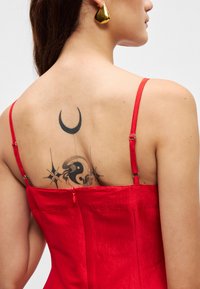 Κόκκινο σατέν φόρεμα με λεπτές τιράντες, που διαθέτει κλείσιμο με φερμουάρ στο πίσω μέρος. Ορατά τατουάζ περιλαμβάνουν μια μισοφέγγαρη και σχέδιο γιν-γιάν.
