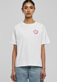 Hvit bomull t-skjorte med korte ermer, med en rosa grafisk logo på venstre brystområde. Løs passform med rund hals.