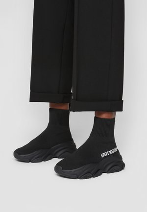 Sorte sokkelignende sneakers med tykke såler og "Steve Madden"-branding, båret med sorte bukser med manchetter på en lys baggrund.