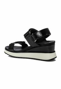 Las sandalias negras de cuña cuentan con dos cintas gruesas, una superficie texturizada y una suela de goma blanca. El diseño es destalonado y tiene correas ajustables en los tobillos.