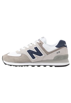 Widok z boku szaro-biało-granatowego buta sportowego New Balance 574 z logo "N" i czarną gumową podeszwą na białym tle.